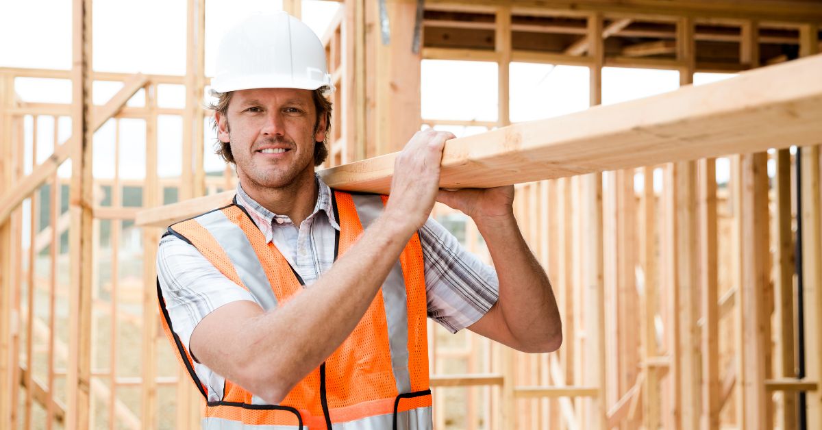 man wearing a hard hat and construction vest carrying a wooden plank in a house foundation